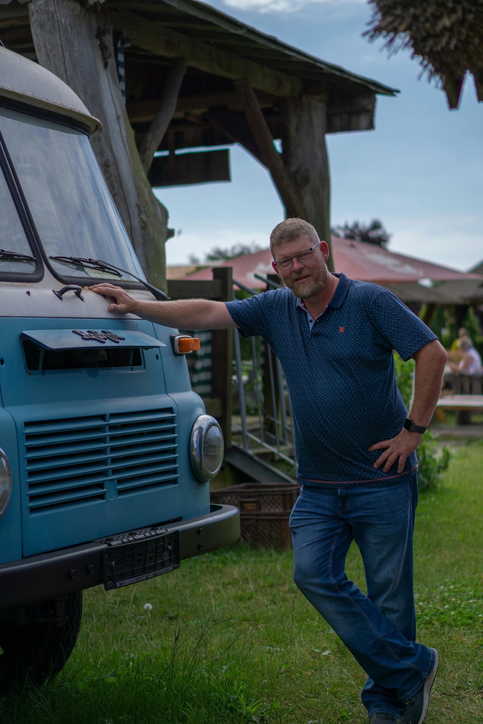 Casual portrait of a man posing with a vintage blue van in a rural setting.